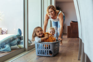 Happy young mother pushing children sitting in laundry basket. Mother and children playing at home.